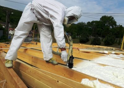 pracovník v bílé kombinéze na střeše aplikující izolační pěnu mezi trámy
