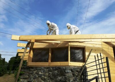 dva pracovníci v bílých kombinézách na střeše dřevěné stavby aplikující izolační pěnu mezi trámy