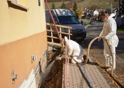 pracovník klečící na chodníku u prohlubně u stěny žlutého domu nanášející izolační materiál